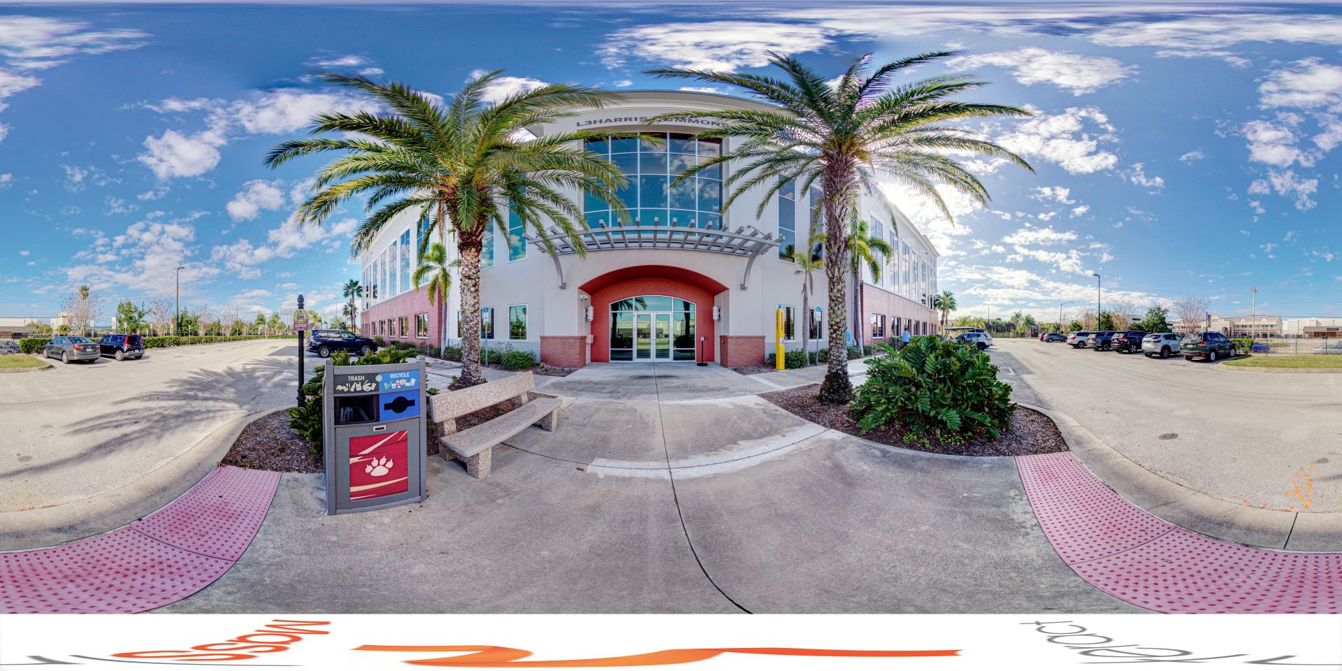 Exterior view of the L3Harris Commons at Florida Tech, featuring the main entrance with a modern facade, palm trees, and a well-maintained parking area under a bright, partly cloudy sky